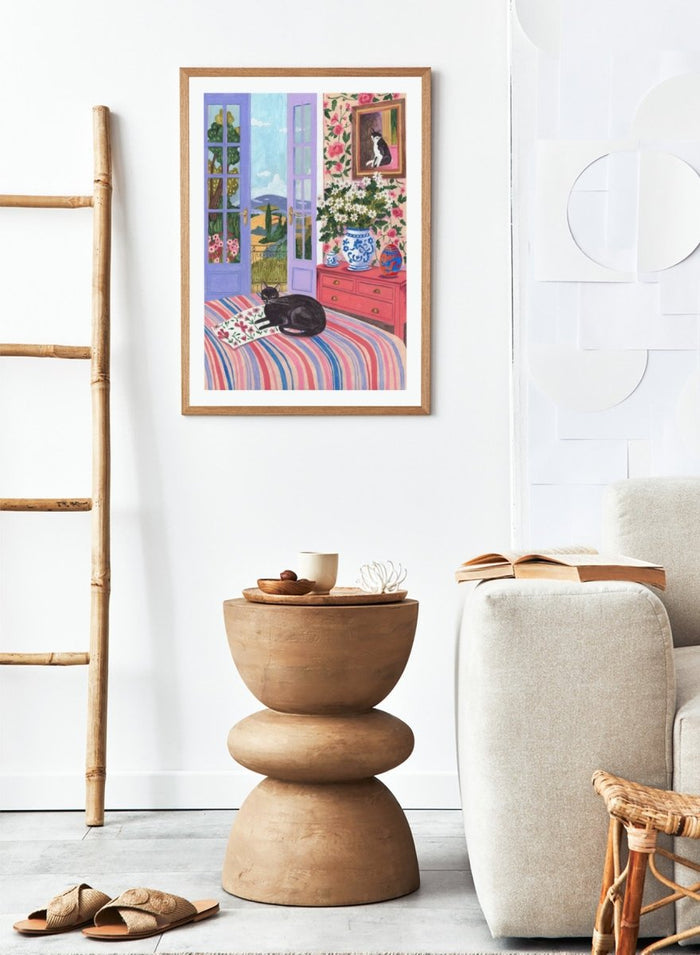 Cat lounging on a striped bed with a view of a garden through open doors, surrounded by flowers and decorative items. Black Cat on Bed French Doors Garden View by Caroline Bonne Müller is a colorful interior art print featuring cat illustration, floral room, and scenic garden view in a modern aesthetic style.