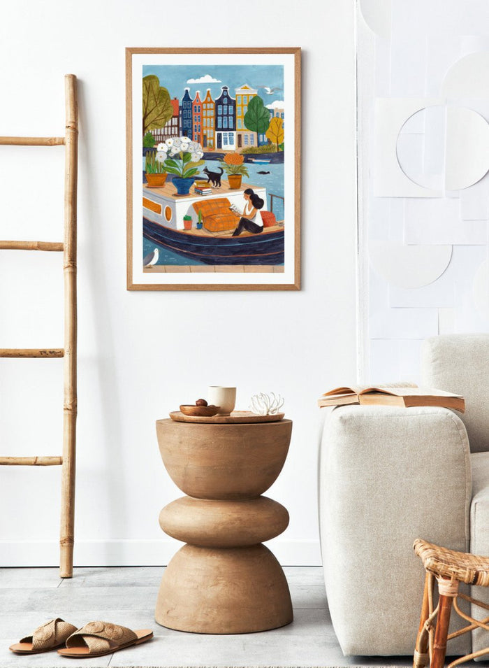 Illustration of a woman on a boat with flowers and a cat, surrounded by colorful houses. Cozy Canal Reading by Caroline Bonne Müller is a fine art print and wall poster featuring a woman reading on a canal boat with colorful European buildings in the background.