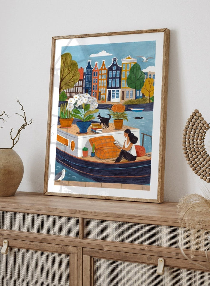 Illustration of a woman on a boat with flowers and a cat, surrounded by colorful houses. Cozy Canal Reading by Caroline Bonne Müller is a fine art print and wall poster featuring a woman reading on a canal boat with colorful European buildings in the background.