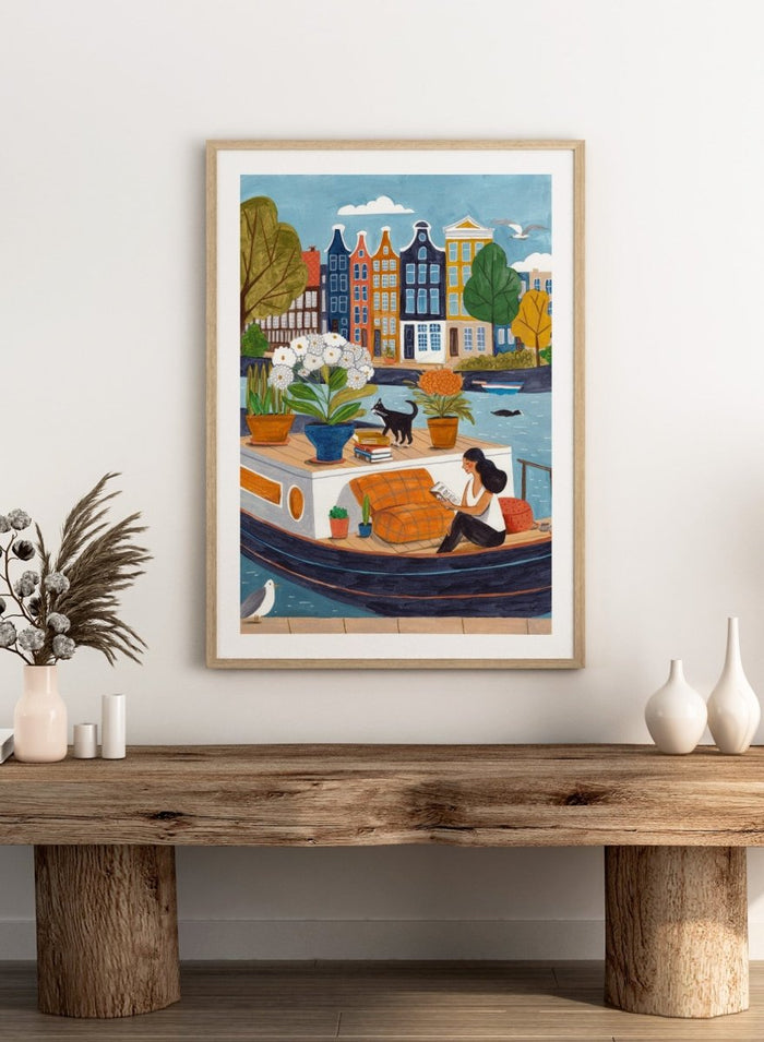 Illustration of a woman on a boat with flowers and a cat, surrounded by colorful houses. Cozy Canal Reading by Caroline Bonne Müller is a fine art print and wall poster featuring a woman reading on a canal boat with colorful European buildings in the background.