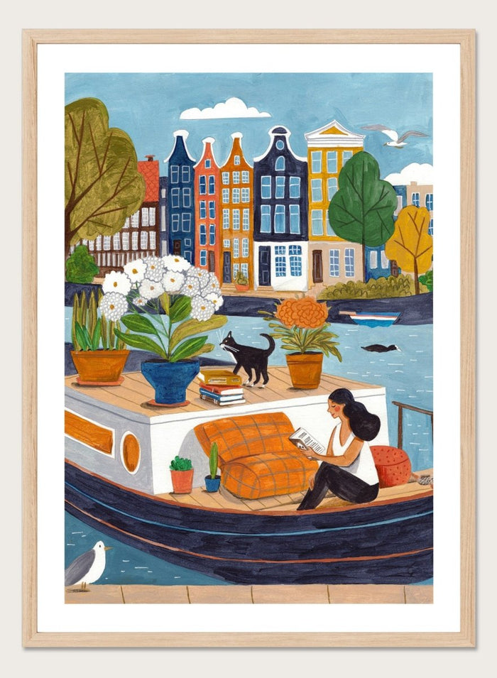 Illustration of a woman on a boat with flowers and a cat, surrounded by colorful houses. Cozy Canal Reading by Caroline Bonne Müller is a fine art print and wall poster featuring a woman reading on a canal boat with colorful European buildings in the background.