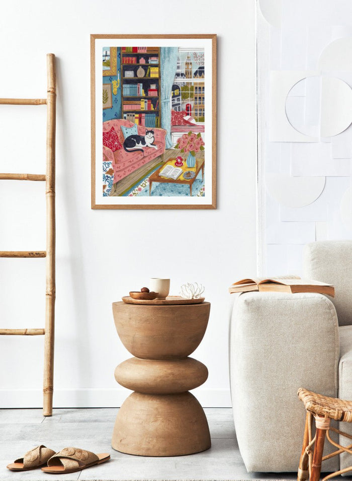 Illustration of a cozy living room with a cat on a pink sofa, bookshelf, and coffee table. Cozy London Cat by Caroline Bonne Müller is a fine art print and wall poster featuring a cat lounging in a colorful living room with a London city view.
