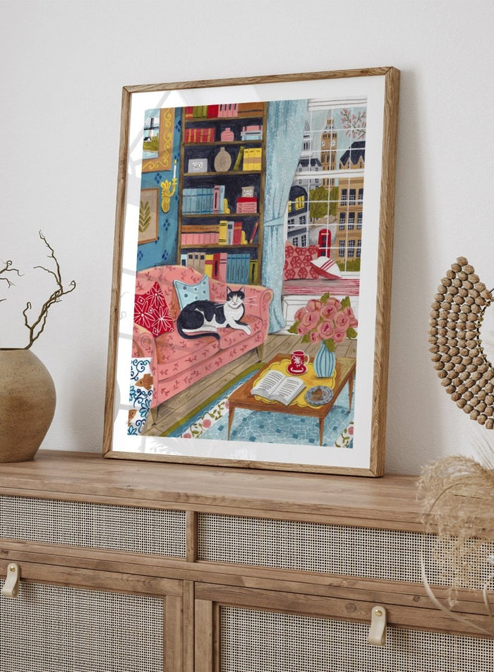 Illustration of a cozy living room with a cat on a pink sofa, bookshelf, and coffee table. Cozy London Cat by Caroline Bonne Müller is a fine art print and wall poster featuring a cat lounging in a colorful living room with a London city view.