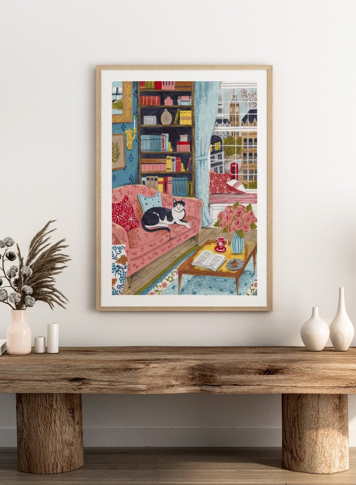 Illustration of a cozy living room with a cat on a pink sofa, bookshelf, and coffee table. Cozy London Cat by Caroline Bonne Müller is a fine art print and wall poster featuring a cat lounging in a colorful living room with a London city view.