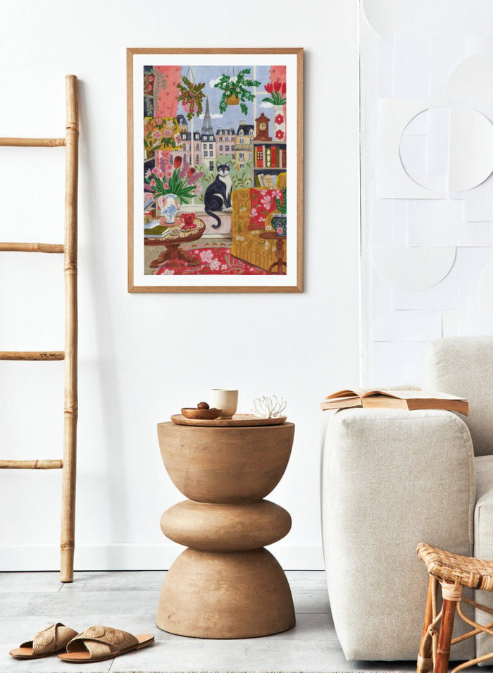 Colorful illustration of a living room with a cat, flowers, and a cityscape. Cozy Parisian Window Cat by Caroline Bonne Müller is a fine art print and wall poster of a cat by a window with Paris view and colorful interior.
