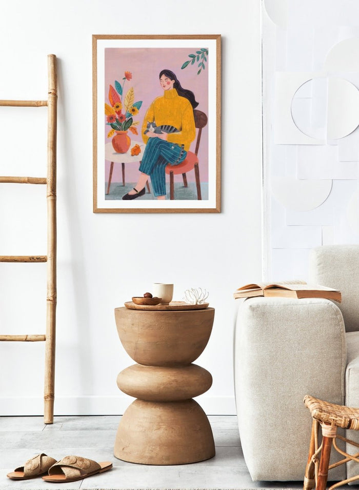 Illustration of a woman in a yellow sweater holding a cat, sitting by a table with flowers on a pink background. Peaceful Woman and Cat by Caroline Bonne Müller is a fine art print and wall poster featuring a woman holding a cat beside a floral arrangement.