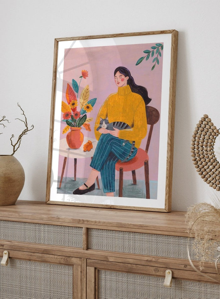 Illustration of a woman in a yellow sweater holding a cat, sitting by a table with flowers on a pink background. Peaceful Woman and Cat by Caroline Bonne Müller is a fine art print and wall poster featuring a woman holding a cat beside a floral arrangement.