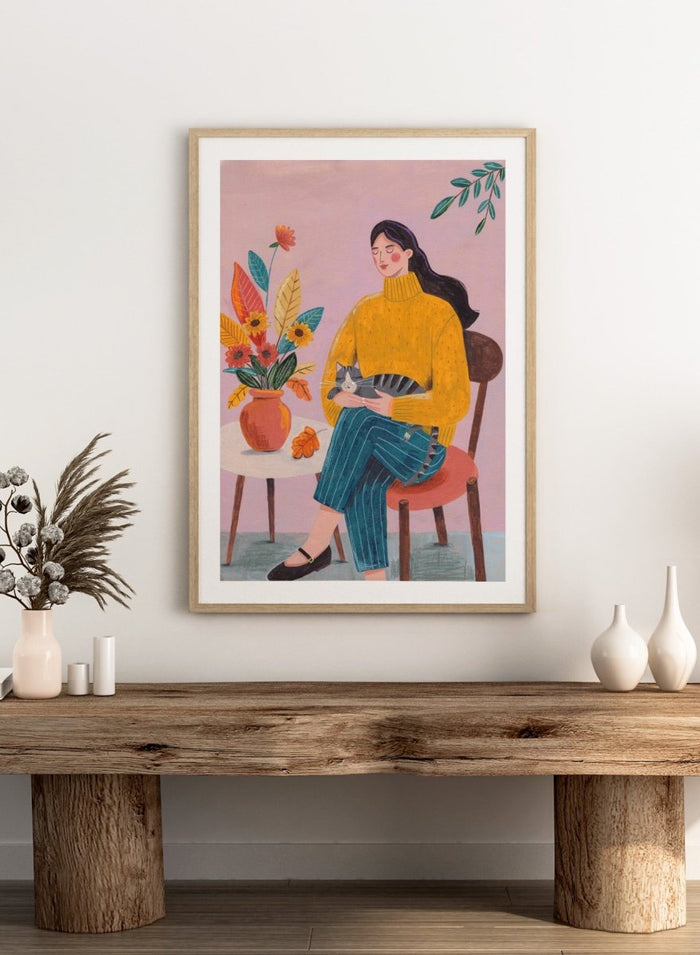Illustration of a woman in a yellow sweater holding a cat, sitting by a table with flowers on a pink background. Peaceful Woman and Cat by Caroline Bonne Müller is a fine art print and wall poster featuring a woman holding a cat beside a floral arrangement.