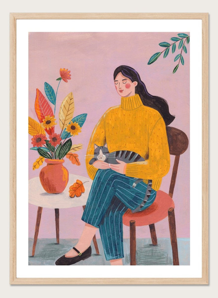 Illustration of a woman in a yellow sweater holding a cat, sitting by a table with flowers on a pink background. Peaceful Woman and Cat by Caroline Bonne Müller is a fine art print and wall poster featuring a woman holding a cat beside a floral arrangement.