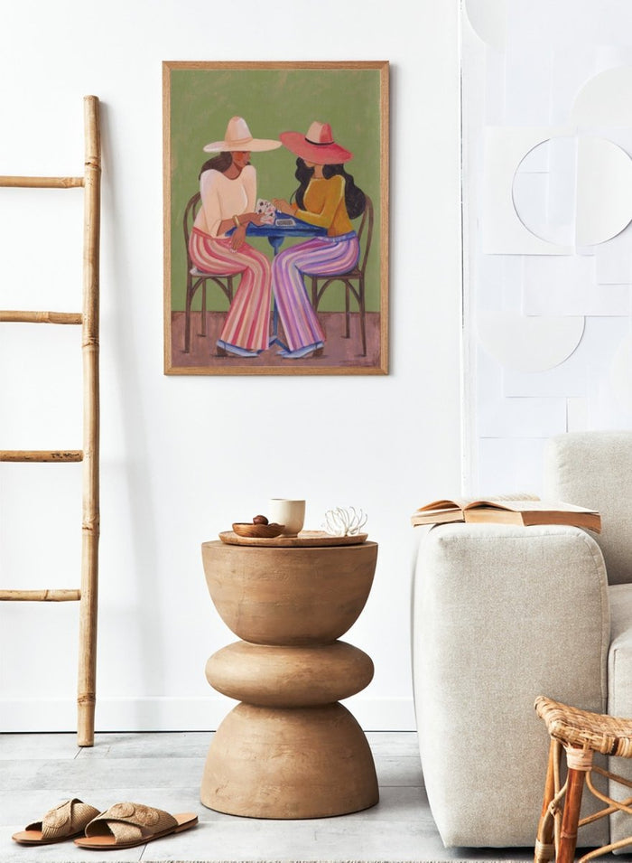 Two women playing cards at a table with colorful clothing and hats against a green background. Stylish Women Play Cards by Caroline Bonne Müller is a fine art print and wall poster featuring two fashionable women playing cards at a table.