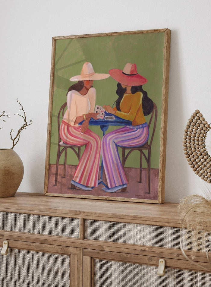 Two women playing cards at a table with colorful clothing and hats against a green background. Stylish Women Play Cards by Caroline Bonne Müller is a fine art print and wall poster featuring two fashionable women playing cards at a table.