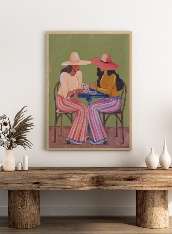 Two women playing cards at a table with colorful clothing and hats against a green background. Stylish Women Play Cards by Caroline Bonne Müller is a fine art print and wall poster featuring two fashionable women playing cards at a table.