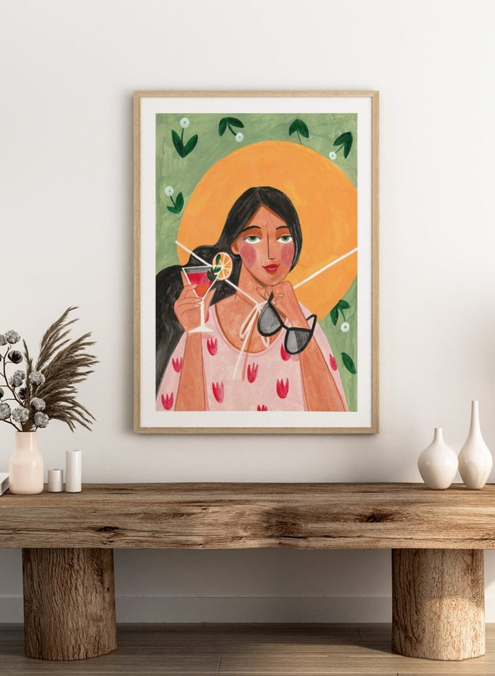 Illustration of a woman holding a cocktail and sunglasses with a sun and foliage background. Summer Woman Cocktail Hat by Caroline Bonne Müller is a fine art print and wall poster featuring a woman holding a cocktail with a bold yellow hat and bright summer colors.
