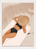 Woman in black bikini and straw hat lying on a towel with sunglasses nearby. Beach Day Relaxation by City Art is a modern beach art print featuring a woman in a black swimsuit, oversized sun hat, soft sand, and warm neutral tones.
