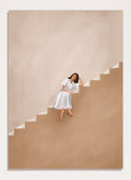 Woman in a white dress walking up a set of stairs with a beige wall and floor. Calm Woman Staircase by City Art is a minimalist figurative art print featuring a woman seated on a staircase with soft neutral tones and modern architectural style.