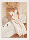 Woman in a white dress holding a clutch, sitting on a couch with a neutral background. Chic Summer Reading by City Art features a woman reading in a sunlit bedroom, bringing warm neutrals, relaxed interiors, and organic summer style home.