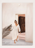 Woman in a white dress standing in a doorway with a palm leaf in the foreground. Woman With Palm Leaf by City Art is a modern figurative art print featuring a woman in a white dress, tropical greenery, warm neutrals, and Mediterranean style.