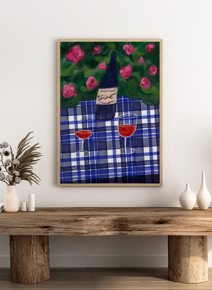 A painted still life scene featuring a dark wine bottle labeled 'vino' on a blue-and-white plaid tablecloth, with two glasses of red wine and pink flowers against a green background. Shop Garden Wine Picnic by Dirkie Nell, a colorful still life art print featuring red wine, blue gingham, pink roses, and a warm summer garden mood.