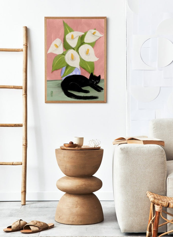 Black cat lying on a surface with white flowers in a blue vase against a pink background. Lilies In a Vase and a Black Cat by Dirkie Nell is a contemporary floral cat art print featuring hand painted lilies, soft pink tones, and playful still life illustration.