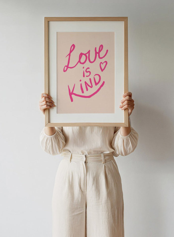 Pink text 'Love is Kind' with a heart on a light pink background. Love Is Kind by Dirkie Nell is a pink typography art print with handwritten lettering and heart detail. A joyful modern love wall poster.