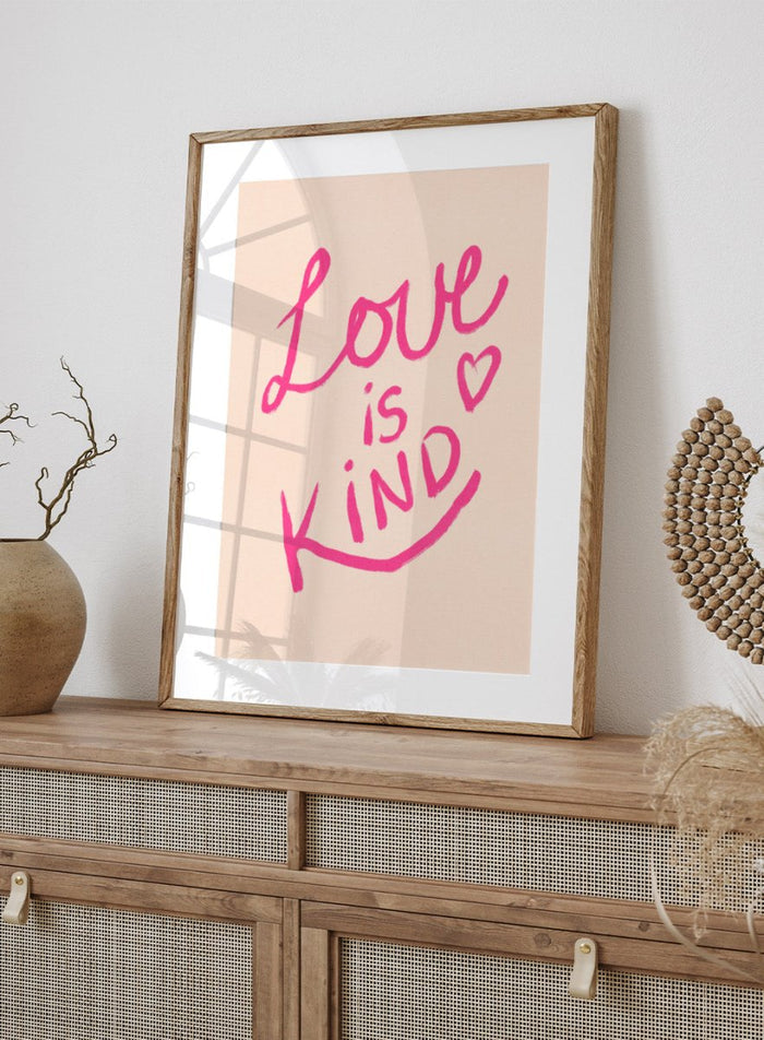 Pink text 'Love is Kind' with a heart on a light pink background. Love Is Kind by Dirkie Nell is a pink typography art print with handwritten lettering and heart detail. A joyful modern love wall poster.