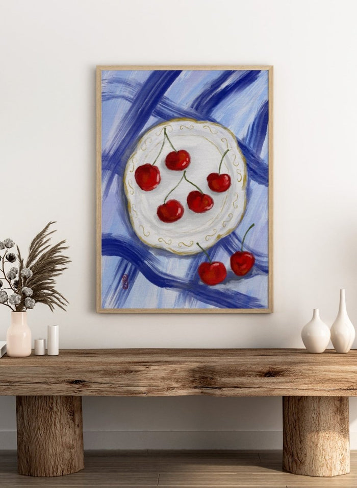 Painting of cherries on a white plate with a blue background. Red Cherries Blue Table by Dirkie Nell is a contemporary fruit still life art print featuring hand painted cherries, blue table linens, and colorful kitchen wall art style.