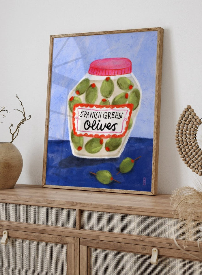 Illustration of a jar of Spanish green olives on a blue background. Spanish Green Olives Jar by Dirkie Nell is a playful food art print featuring hand painted olives, colorful kitchen details, and Mediterranean inspired design.