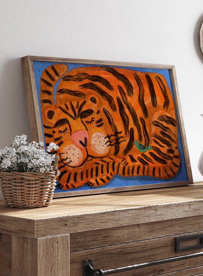 A framed painting of a sleeping tiger on a blue background. The tiger is orange with bold black stripes, has a pink nose and cheeks, and a small green bird near its side. A framed painting of a sleeping tiger on a blue background. The tiger is orange with bold black stripes, has a pink nose and cheeks, and a small green bird near its side.