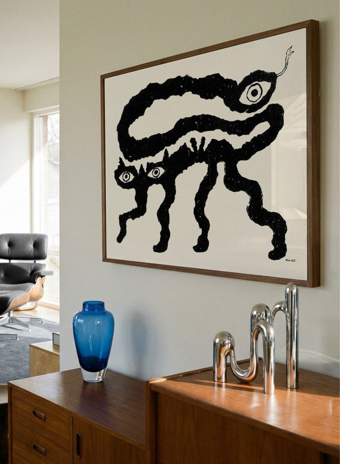 Framed artwork of a black and white creature with cat-like features and snake-like body. I Am Animals by Eden Kalif is a modern minimalist line art print featuring abstract animal forms in a bold black and white contemporary wall poster design.