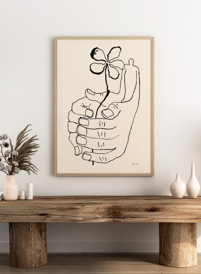 Black line drawing of a hand holding a flower on a beige background. Let It Grow by Eden Kalif is a modern line art print featuring a hand holding a flower with a human figure, designed as a minimalist contemporary wall poster.