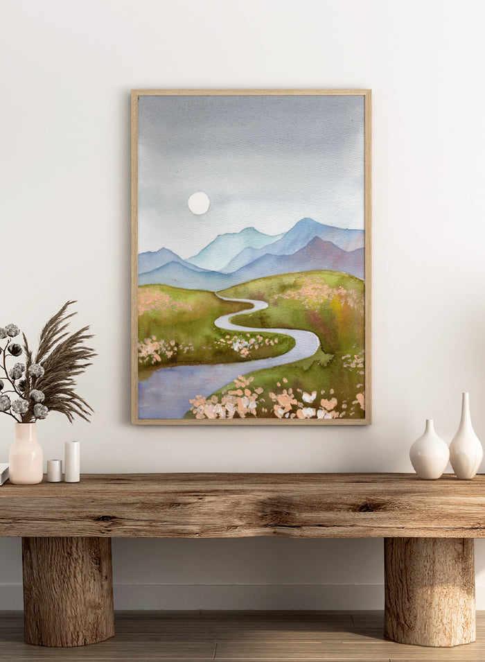 Watercolor landscape painting of a winding river flowing through green hills with pink flowers, blue mountains in the background, and a white sun in a gray sky. Shop Serene Mountain Path by Ekaterina Prisich, a landscape art print featuring a winding valley path, blue mountains, soft flowers, and a white moon.