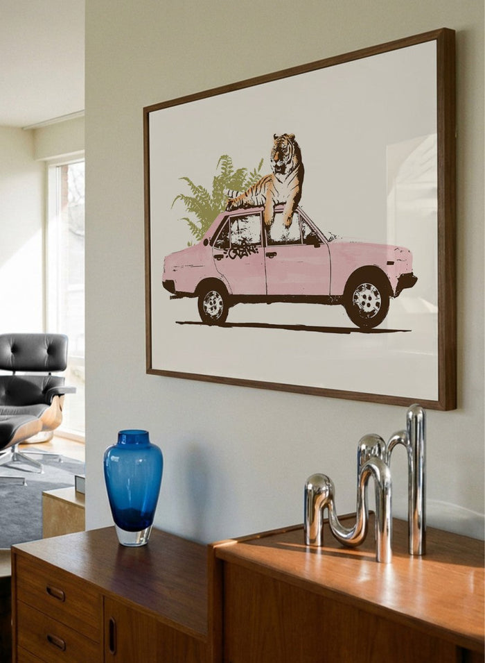 Framed artwork of a tiger in a pink car with greenery on a white background. Tiger On Pink Car by Florent Bodart is a surreal animal art print with a tiger on a vintage car, a modern wall poster with playful, bold, and imaginative design.