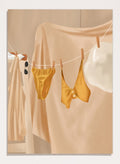 Yellow bikini top and bottom hanging on a clothesline against a beige background. Golden Summer Laundry by Flow Line is a summer art print featuring hanging linens, a yellow swimsuit, sun hat, and Mediterranean-inspired coastal decor.