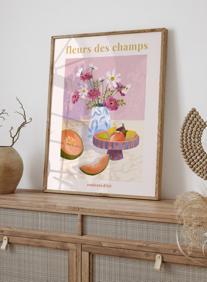 Illustration of a vase with flowers and fruits on a table against a pink background, with text 'fleurs des champs' and 'couleurs d'ete'. Fleur Des Champs Pink by Goed Blauw is a colorful still life art print and wall poster featuring flowers, fruit, and a patterned vase in a modern composition.