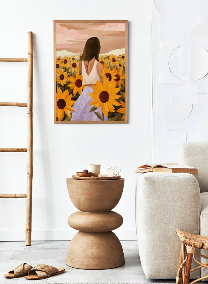 Woman in a white top and purple pants sitting among sunflowers with a scenic background. Sunflower Evenings by Goed Blauw is a fine art print and wall poster featuring a woman standing in a sunflower field with warm golden tones.