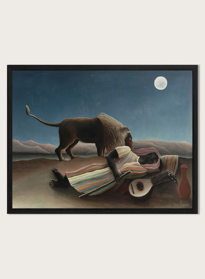 A framed surreal art print depicting a lion standing over a sleeping person wrapped in a striped blanket, with a guitar, vase, and full moon in a night sky. The Sleeping Gypsy by Henri Rousseau is a vintage art print featuring a sleeping traveler, lion, moonlit desert, striped robe, mandolin, and deep blue night sky.