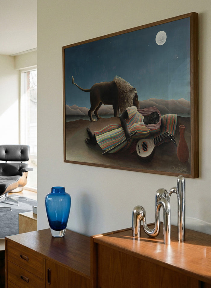 A framed surreal art print depicting a lion standing over a sleeping person wrapped in a striped blanket, with a guitar, vase, and full moon in a night sky. The Sleeping Gypsy by Henri Rousseau is a vintage art print featuring a sleeping traveler, lion, moonlit desert, striped robe, mandolin, and deep blue night sky.