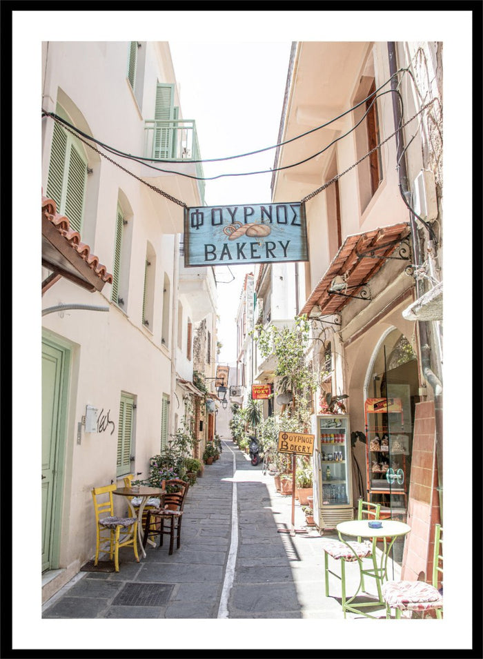narrow street with a bakery sign in a European town. Charming Bakery Street by Henrike Schenk is a Greek street photography print featuring a Mediterranean bakery alley, pastel buildings, café seating, and travel wall art.
