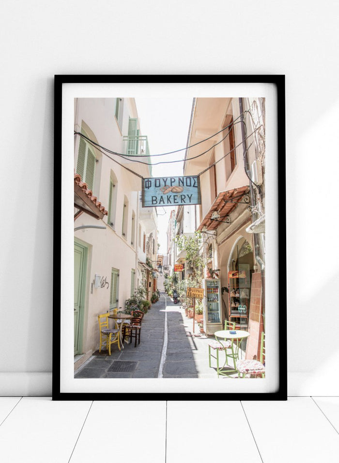 narrow street with a bakery sign in a European town. Charming Bakery Street by Henrike Schenk is a Greek street photography print featuring a Mediterranean bakery alley, pastel buildings, café seating, and travel wall art.