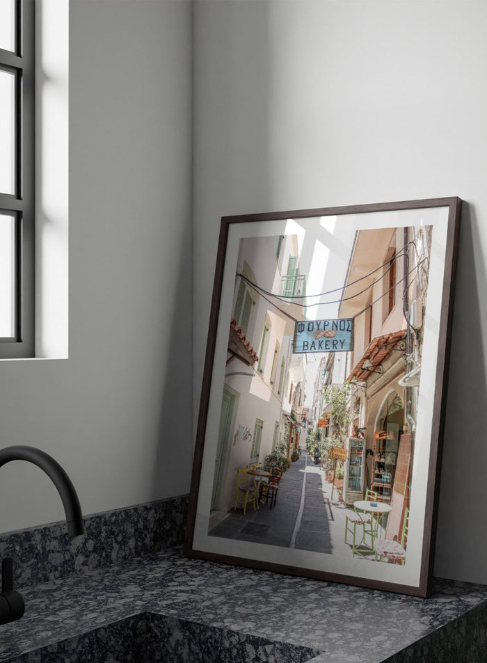 narrow street with a bakery sign in a European town. Charming Bakery Street by Henrike Schenk is a Greek street photography print featuring a Mediterranean bakery alley, pastel buildings, café seating, and travel wall art.