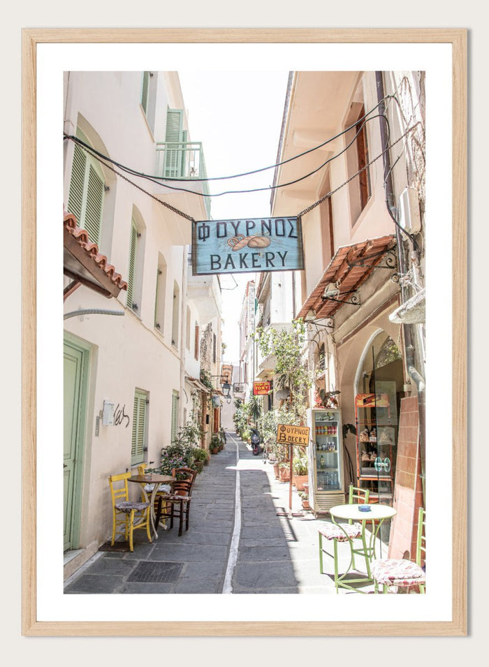 narrow street with a bakery sign in a European town. Charming Bakery Street by Henrike Schenk is a Greek street photography print featuring a Mediterranean bakery alley, pastel buildings, café seating, and travel wall art.