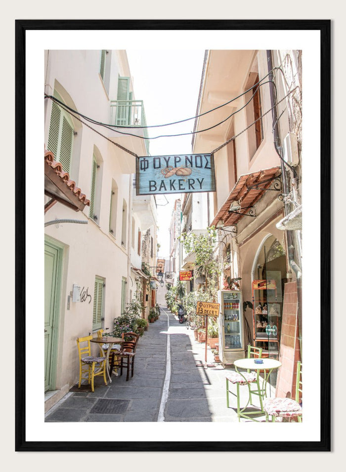 narrow street with a bakery sign in a European town. Charming Bakery Street by Henrike Schenk is a Greek street photography print featuring a Mediterranean bakery alley, pastel buildings, café seating, and travel wall art.