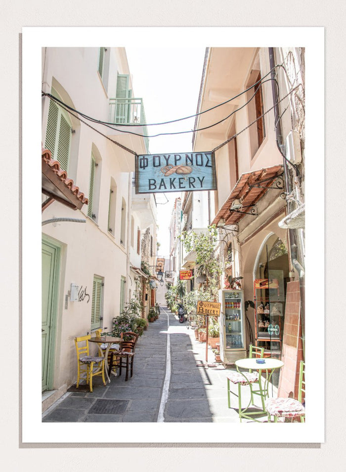 narrow street with a bakery sign in a European town. Charming Bakery Street by Henrike Schenk is a Greek street photography print featuring a Mediterranean bakery alley, pastel buildings, café seating, and travel wall art.