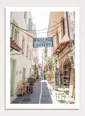 narrow street with a bakery sign in a European town. Charming Bakery Street by Henrike Schenk is a Greek street photography print featuring a Mediterranean bakery alley, pastel buildings, café seating, and travel wall art.