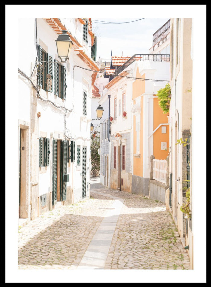 Cobbled street in a quaint European town with white buildings and a clear sky. Charming European Cobblestone Street by Henrike Schenk is a travel photography print featuring cobblestone streets, historic architecture, and European wall art.