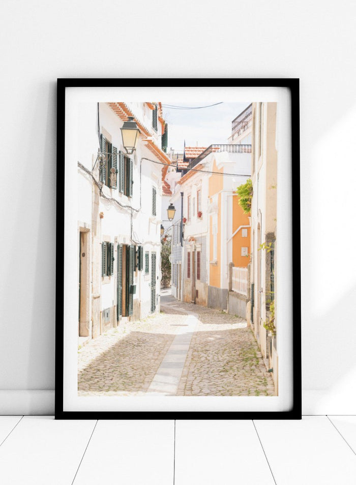 Cobbled street in a quaint European town with white buildings and a clear sky. Charming European Cobblestone Street by Henrike Schenk is a travel photography print featuring cobblestone streets, historic architecture, and European wall art.