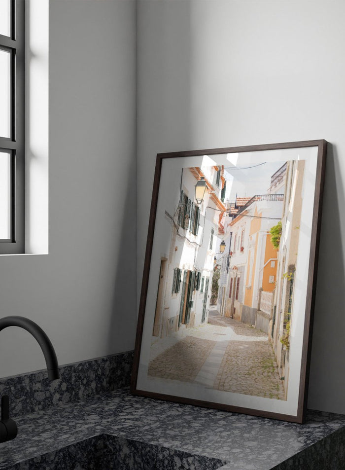 Cobbled street in a quaint European town with white buildings and a clear sky. Charming European Cobblestone Street by Henrike Schenk is a travel photography print featuring cobblestone streets, historic architecture, and European wall art.