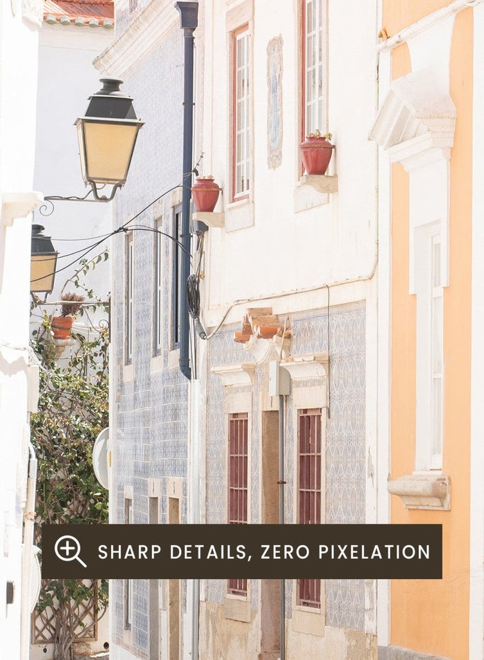 Cobbled street in a quaint European town with white buildings and a clear sky. Charming European Cobblestone Street by Henrike Schenk is a travel photography print featuring cobblestone streets, historic architecture, and European wall art.