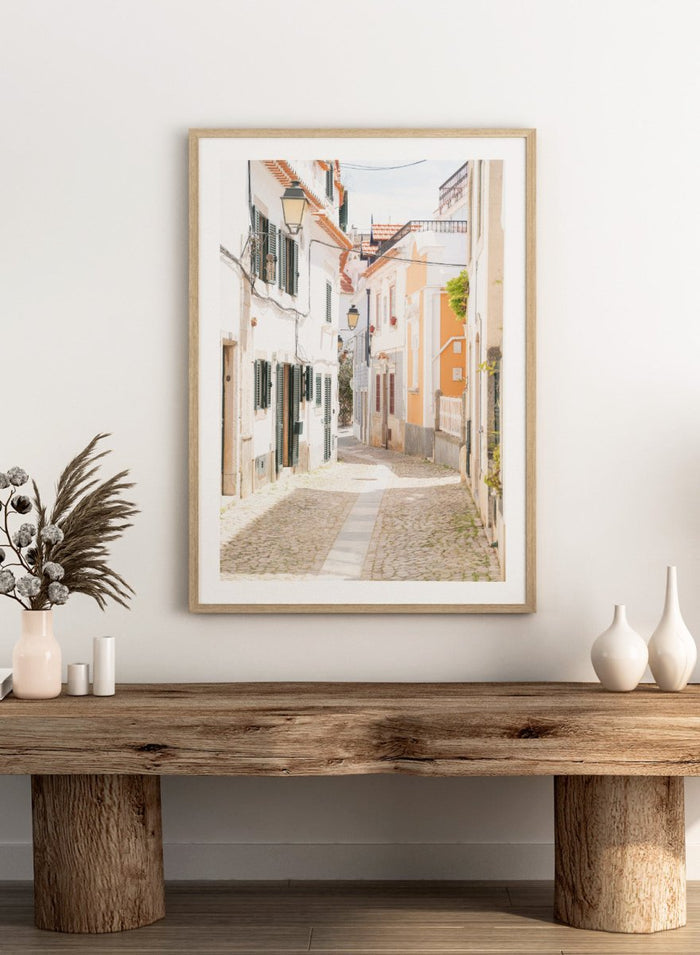 Cobbled street in a quaint European town with white buildings and a clear sky. Charming European Cobblestone Street by Henrike Schenk is a travel photography print featuring cobblestone streets, historic architecture, and European wall art.
