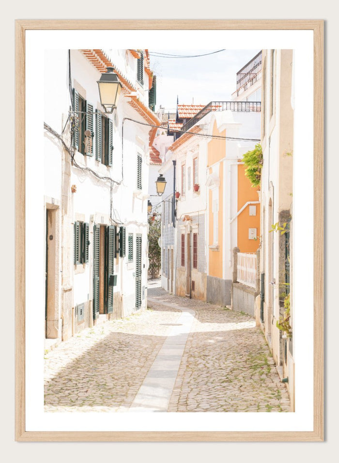 Cobbled street in a quaint European town with white buildings and a clear sky. Charming European Cobblestone Street by Henrike Schenk is a travel photography print featuring cobblestone streets, historic architecture, and European wall art.